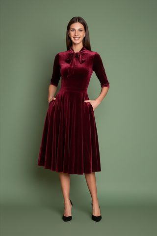Μπορντό φόρεμα, εμπνευσμένο από τη λάμψη της δεκαετίας του 1950, από πολυτελές απαλό βελούδο, με κολακευτική λαιμόκοψη V που τονίζεται από έναν γοητευτικό φιόγκο από ύφασμα που δένει μπροστά. Οι ώμοι με την ελαφριά σούρα και τα 3/4 μανίκια προσθέτουν μια vintage πινελιά, ενώ η σταθερή ζώνη αγκαλιάζει τη σιλουέτα για ένα θηλυκό σχήμα. Σχεδιασμένο με κρυφές τσέπες με πλαϊνές ραφές και φούστα σε κλος γραμμή που κινείται με χάρη σε κάθε βήμα. Ιδανικό για επίσημες περιστάσεις ή απλώς για να αγκαλιάσει το κλασικό στυλ με μια μοντέρνα πινελιά.